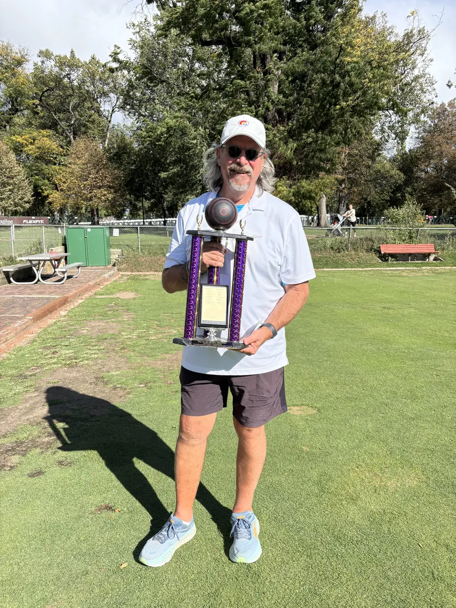 A 2025 club champion in a white cap and white shirt holds the purple championship trophy on the sunny bowling green, trees and a red bench in the background