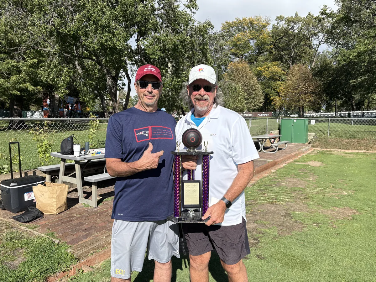 2025 club champions: two smiling bowlers in ball caps and sunglasses stand on the green at Washington Park Lawn Bowling Club, one giving a thumbs-up, holding a tall purple trophy topped with a bowl, with trees and the clubhouse fence behind