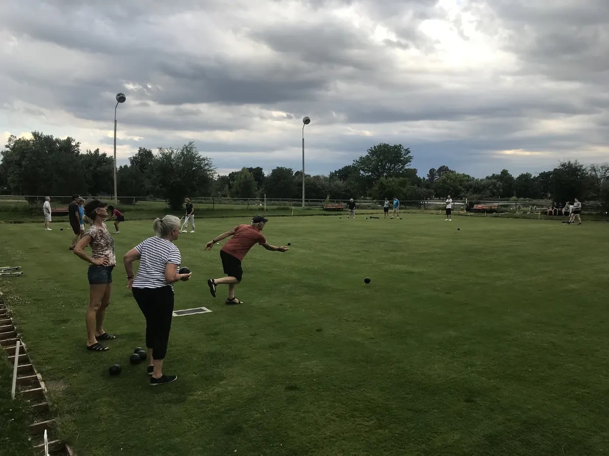 A bowler in a red shirt lunges forward to deliver a bowl while two women in the foreground wait their turn, under a moody cloudy sky with park light poles and trees behind