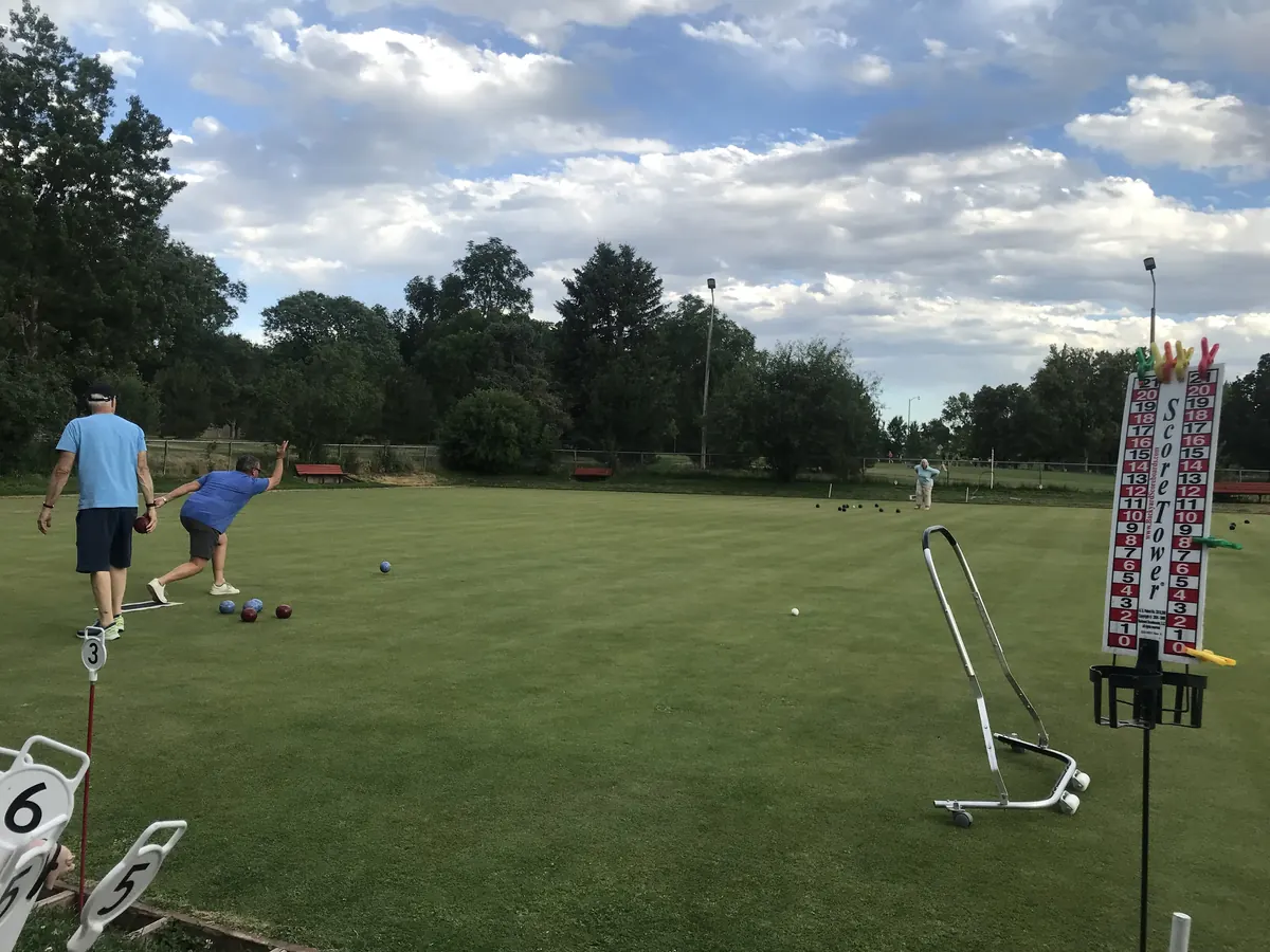 A bowler in a blue shirt follows through on a delivery as a teammate looks on, with a Score Tower scoreboard and rink number markers at the edge of the green